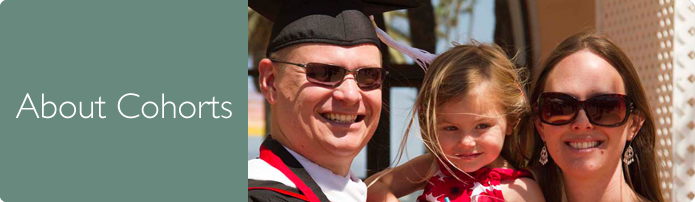 About Cohorts. Smiling student in graduation cap and gown standing next to his wife and holding his daugter.