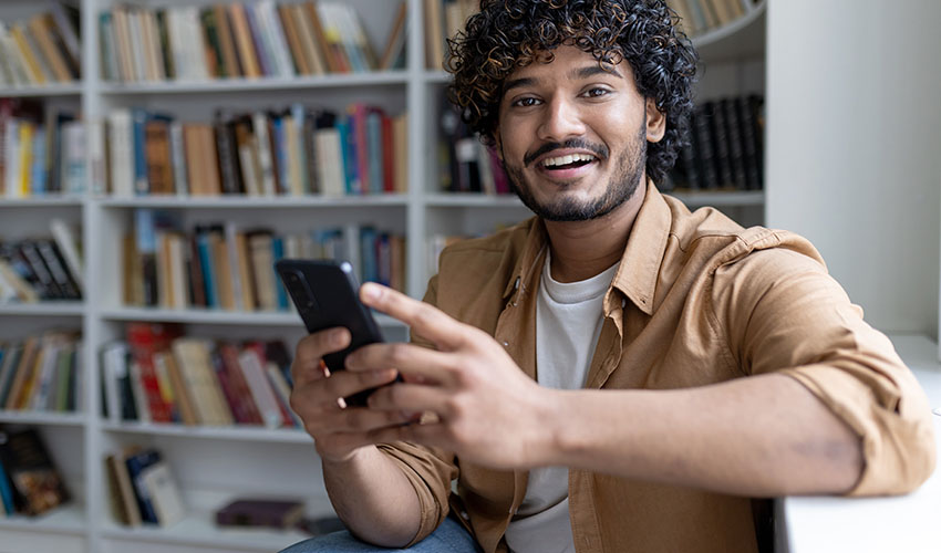 A smiling man with curly hair holds a cell phone, representing the transition to digital student ID cards.