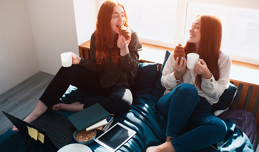 Two women on a bed enjoy coffee and croissants as they study together.