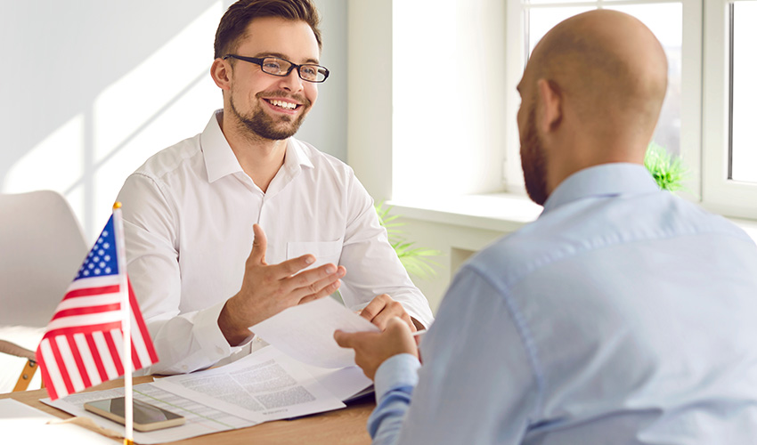 A man wearing glasses engages in conversation with another man at a desk, both focused and attentive.