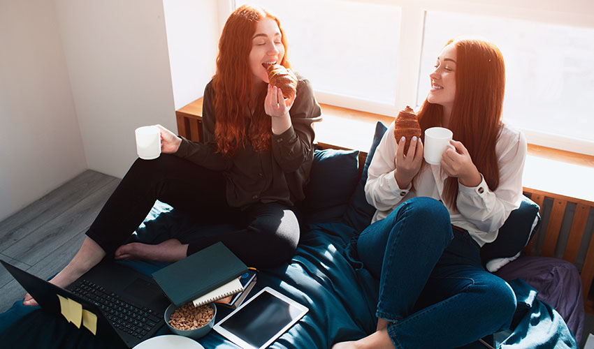 Two women on a bed enjoy coffee and croissants as they study together.