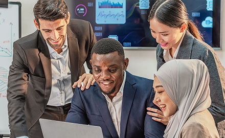 A group of students in business attire collaborates around a laptop, engaged in a lively discussion.